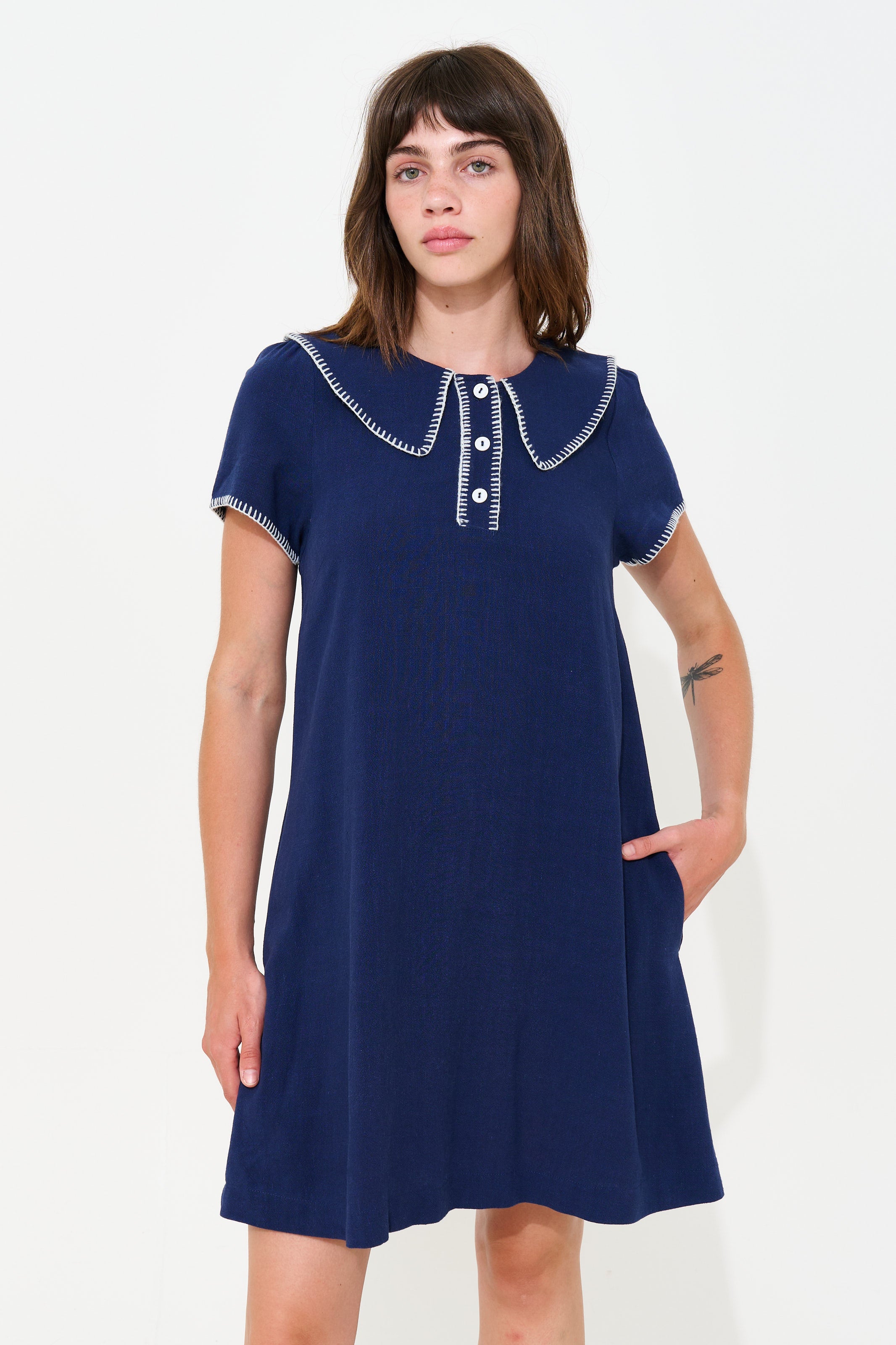 A woman modeling a navy blue short-sleeve dress with a decorative white embroidered collar and button placket, set against a plain white studio background.