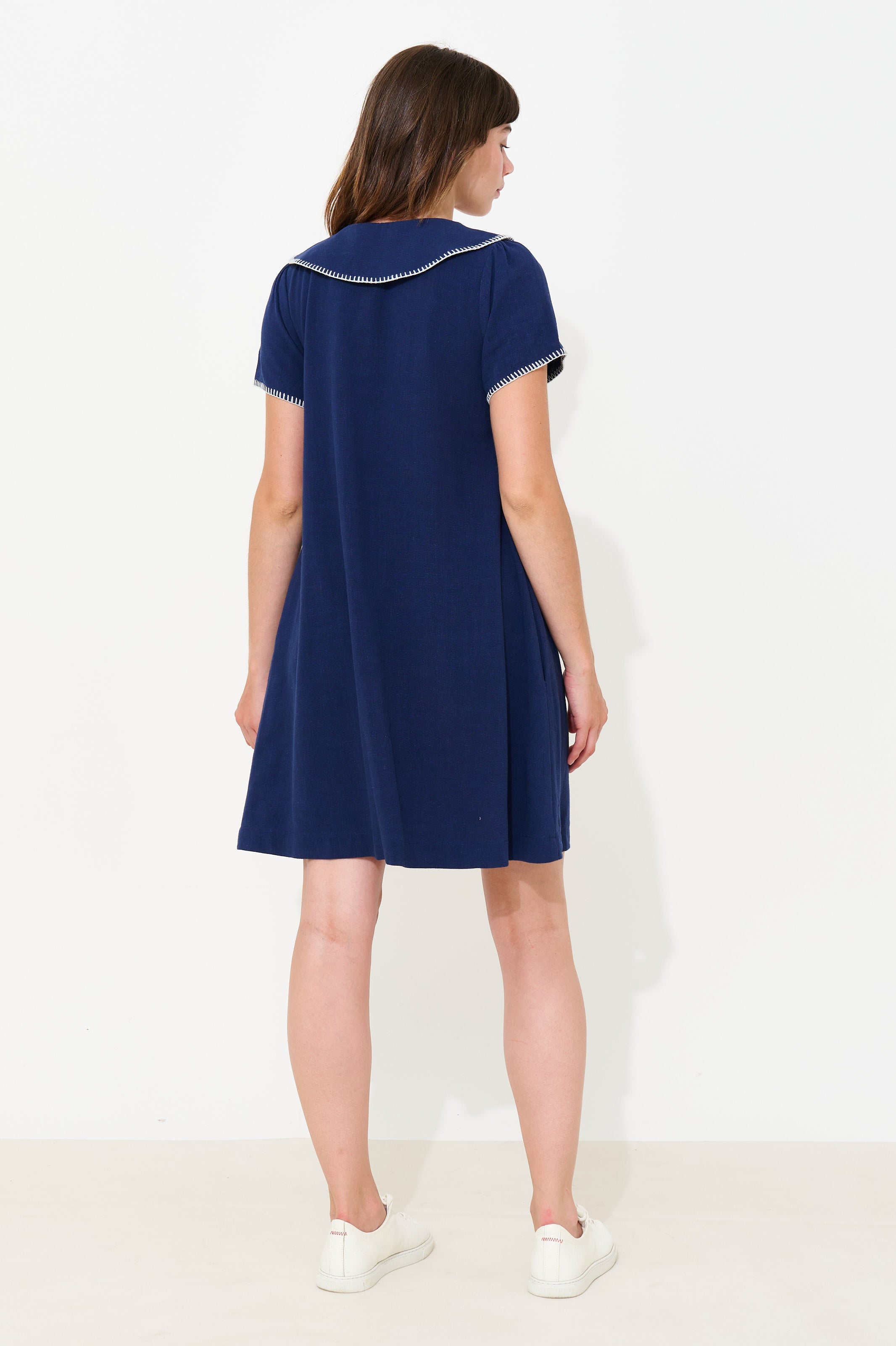 A woman shown from the back wearing a navy blue short-sleeve dress with white trim along the collar and sleeve edges, paired with white sneakers, against a plain white studio background.