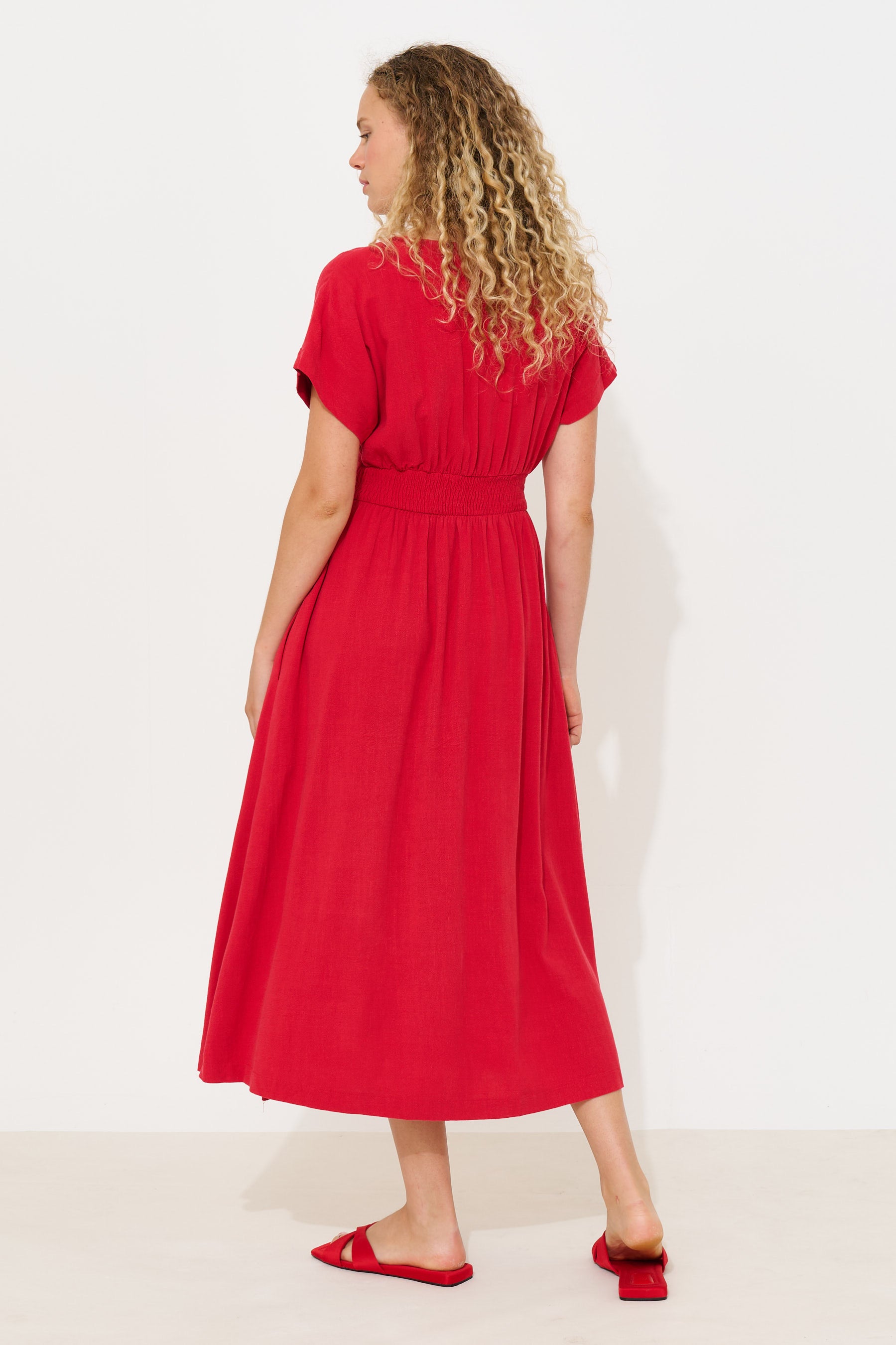 A woman with curly blonde hair is shown from the back wearing a bright red midi dress with short sleeves and a gathered waist, paired with matching red flat sandals, against a plain white studio background.