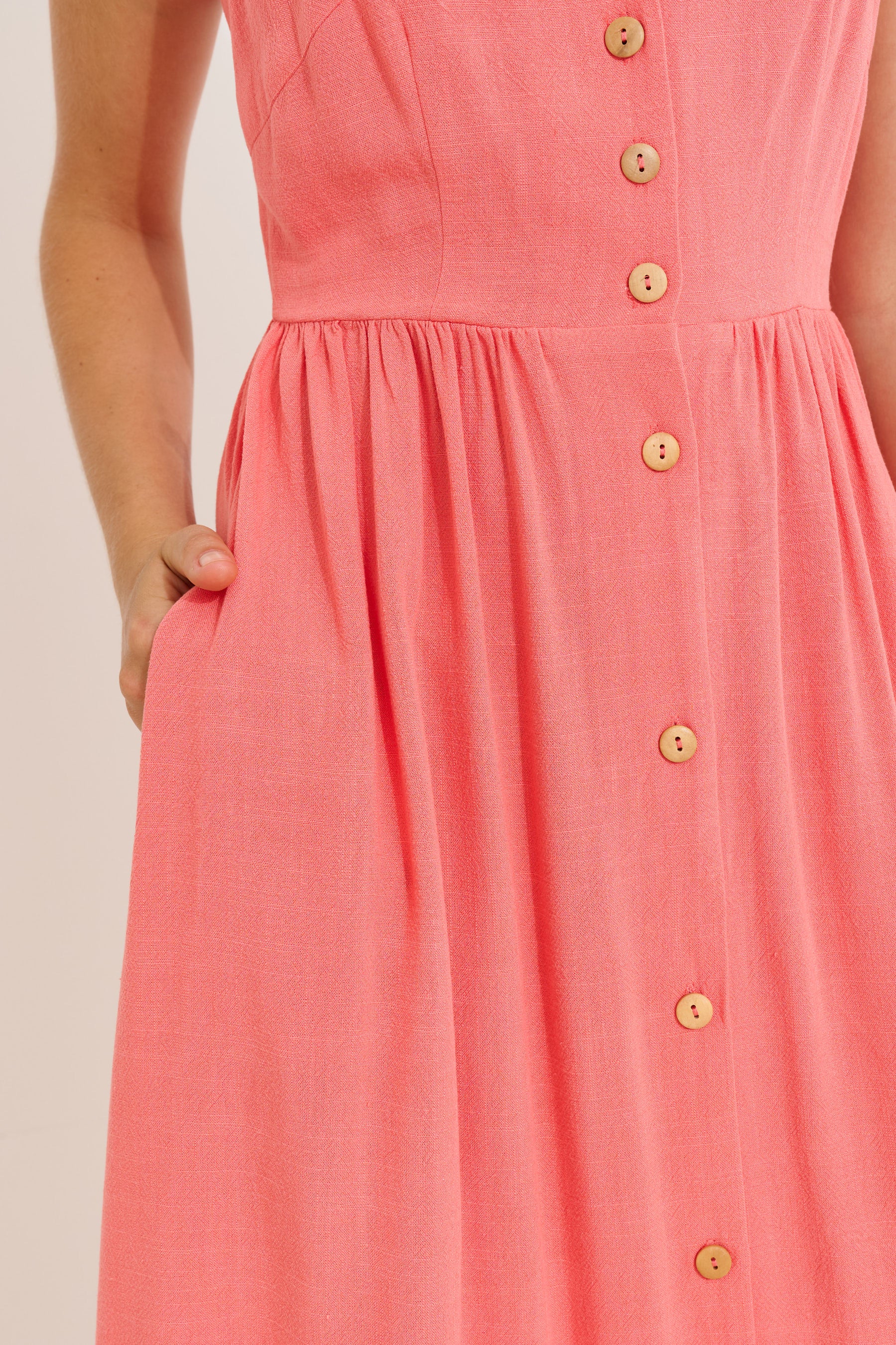Close-up of a coral pink sleeveless dress with a gathered waist, front button closure, and side pockets, shown on a model against a light neutral background.