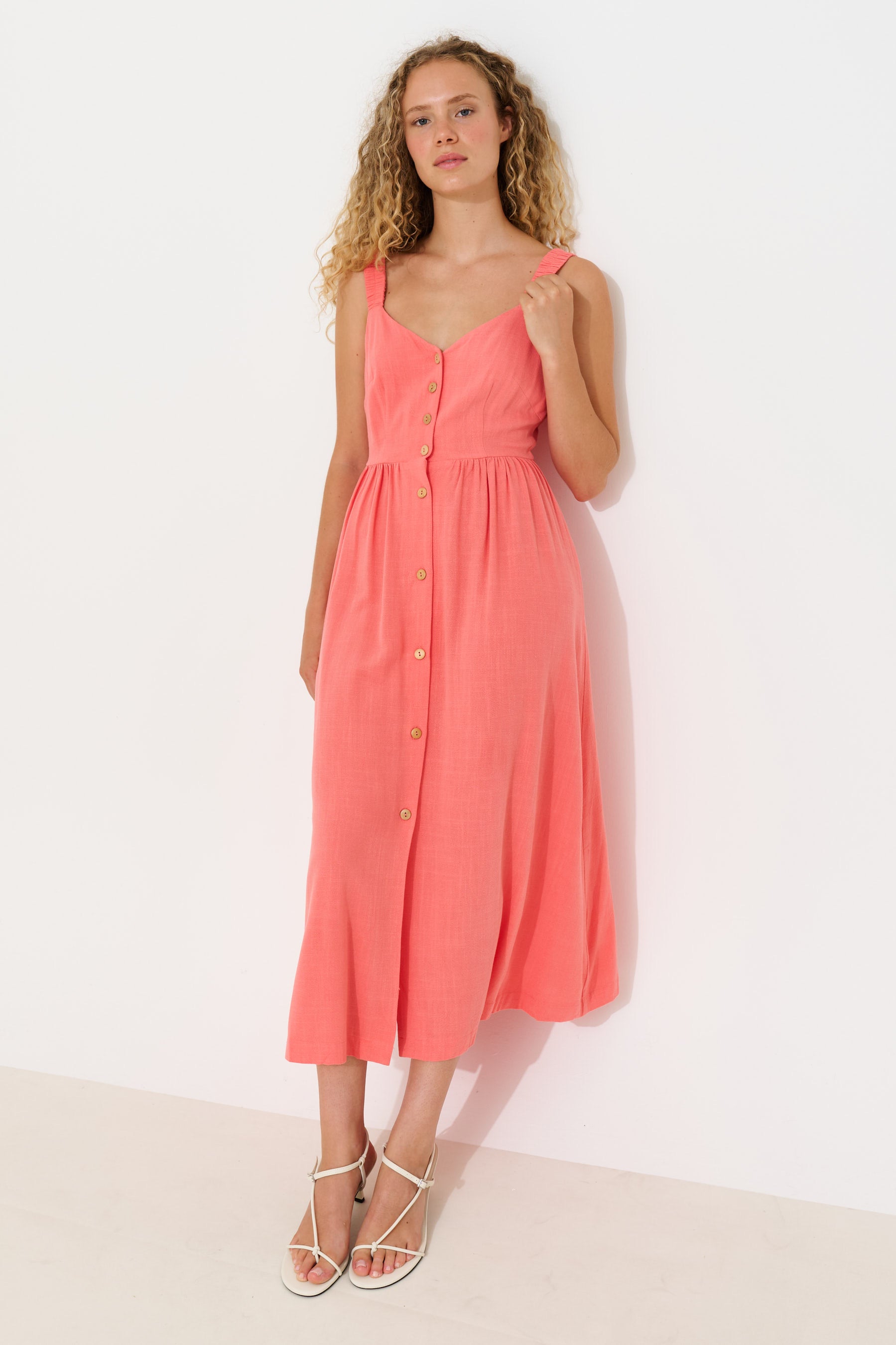 A woman modeling a sleeveless coral pink midi dress with a button-down front, standing against a plain white wall.