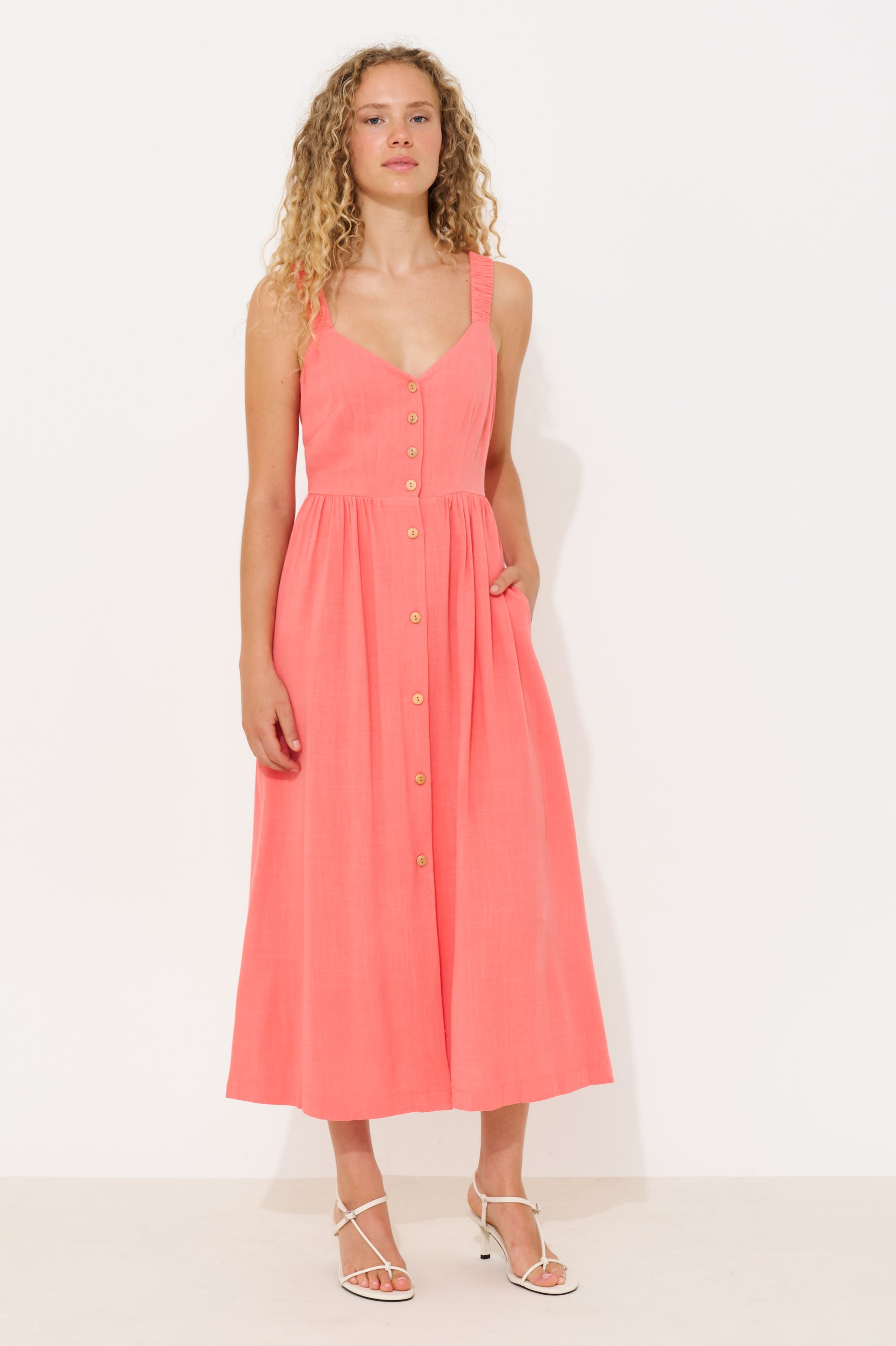 A woman modeling a sleeveless coral pink midi dress with a button-down front, V-neckline, and side pockets, paired with white strappy heels against a plain white studio background.