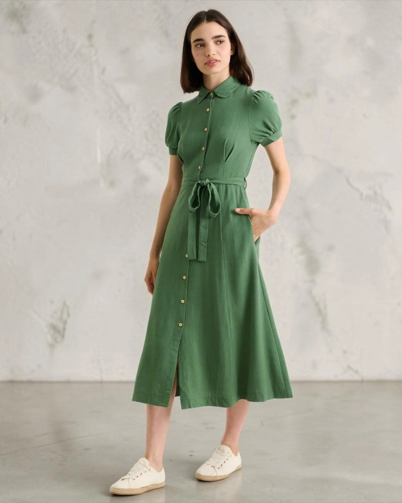 A woman stands against a neutral textured background wearing a green short-sleeve button-front midi dress with a tie waist and white trainers.