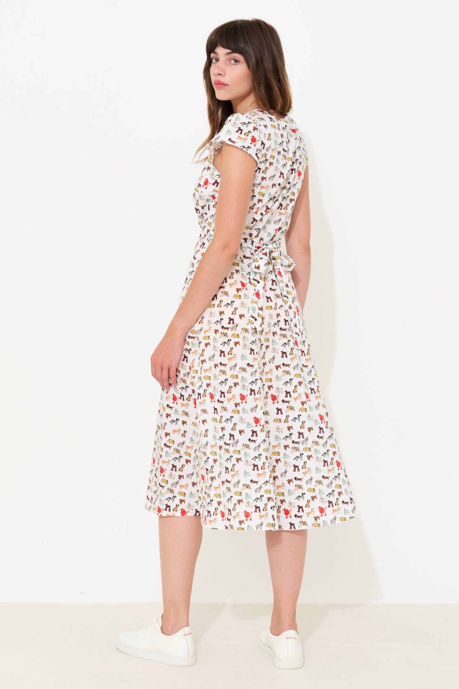 A woman modeling a white knee-length dress with a colorful all-over print, shown from a side-back angle against a plain white studio background.
