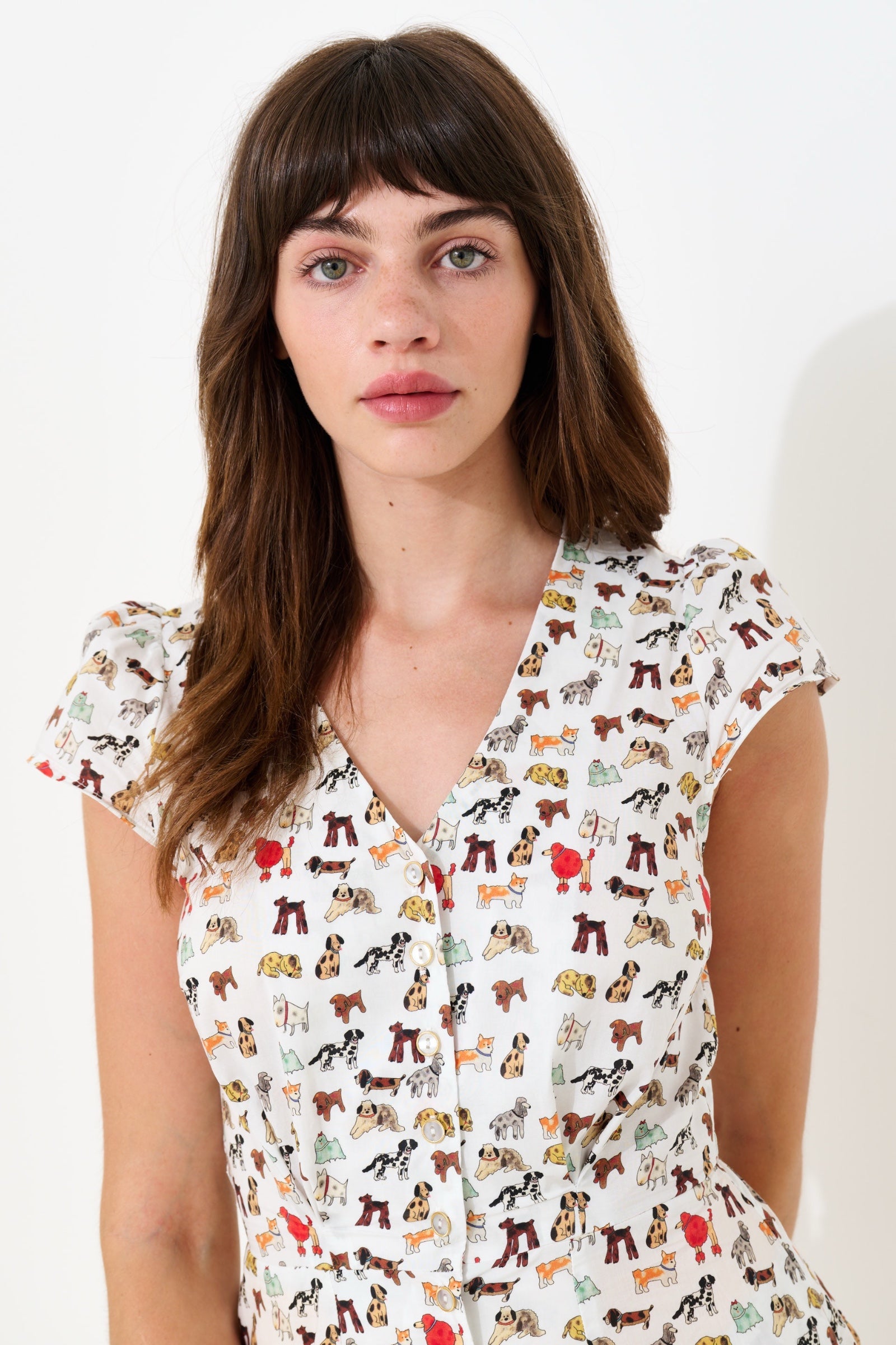 A woman with brown hair and bangs wearing a white short-sleeve button-up dress with a colorful cartoon dog print, photographed against a plain white background.