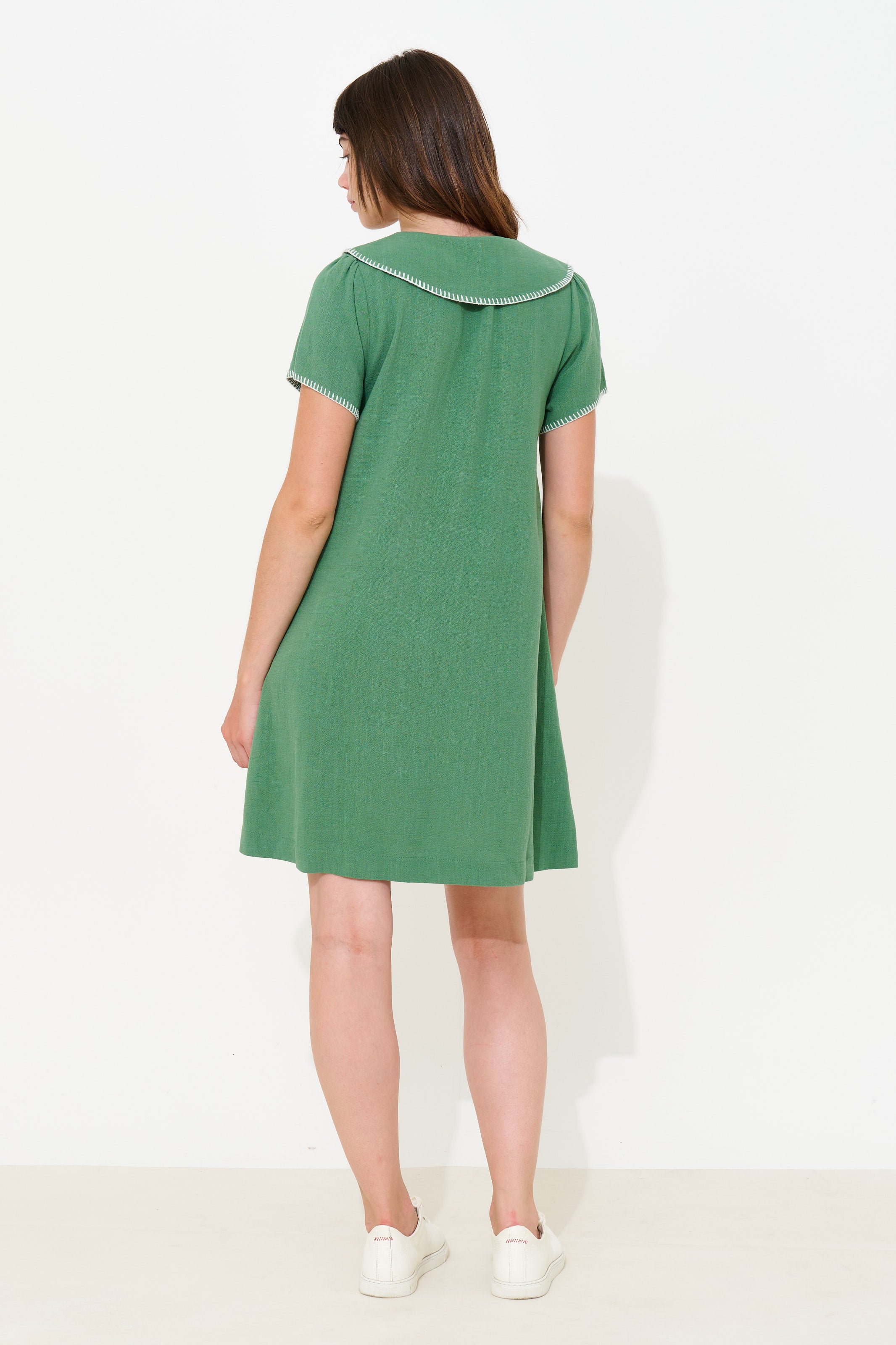 A woman viewed from the back wearing a green short-sleeve dress with a white collar trim, paired with white sneakers, against a plain white studio background.