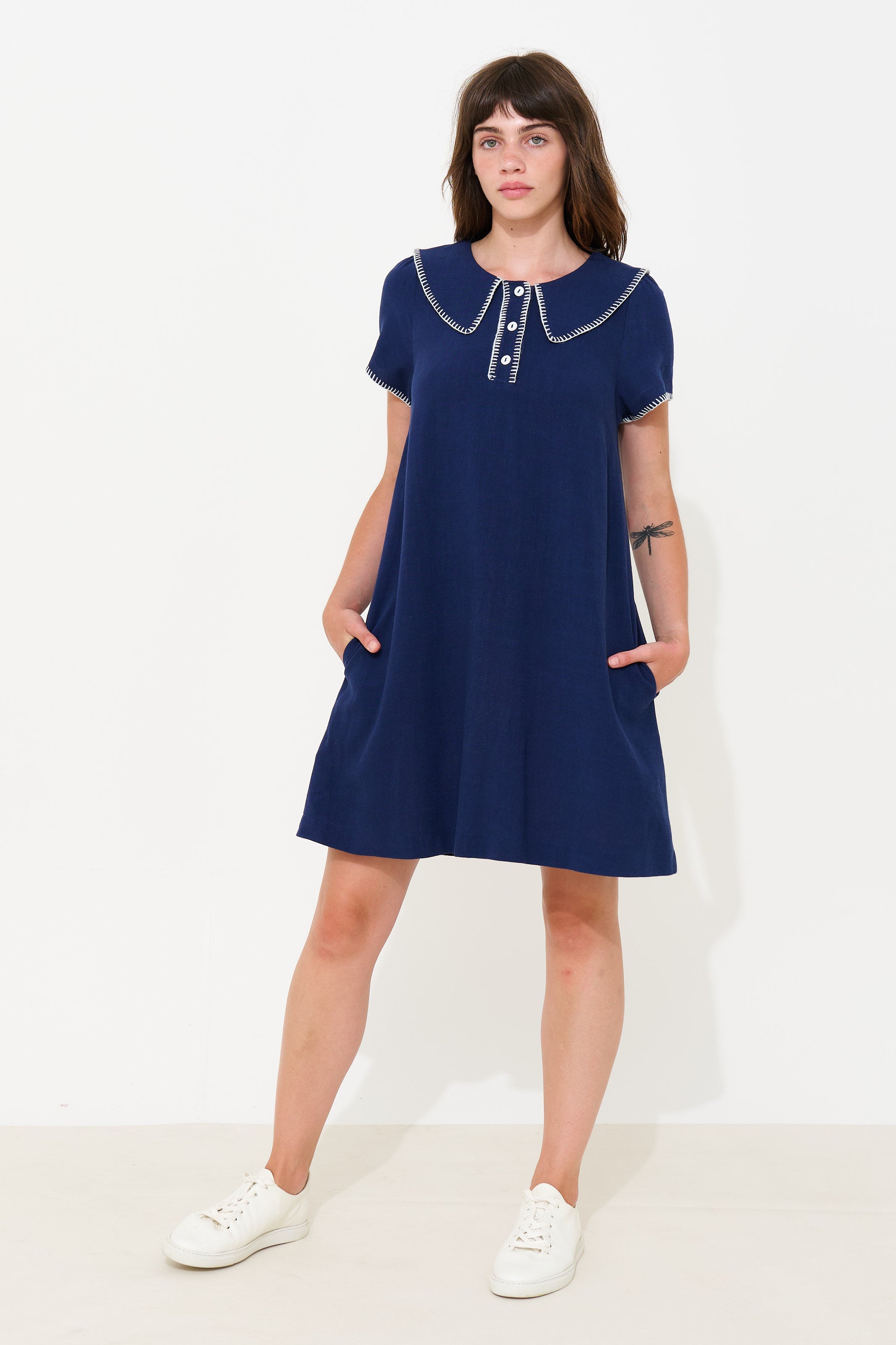 A woman modeling a navy blue short-sleeve shift dress with a Peter Pan collar and white button accents, paired with white sneakers, against a plain white studio background.