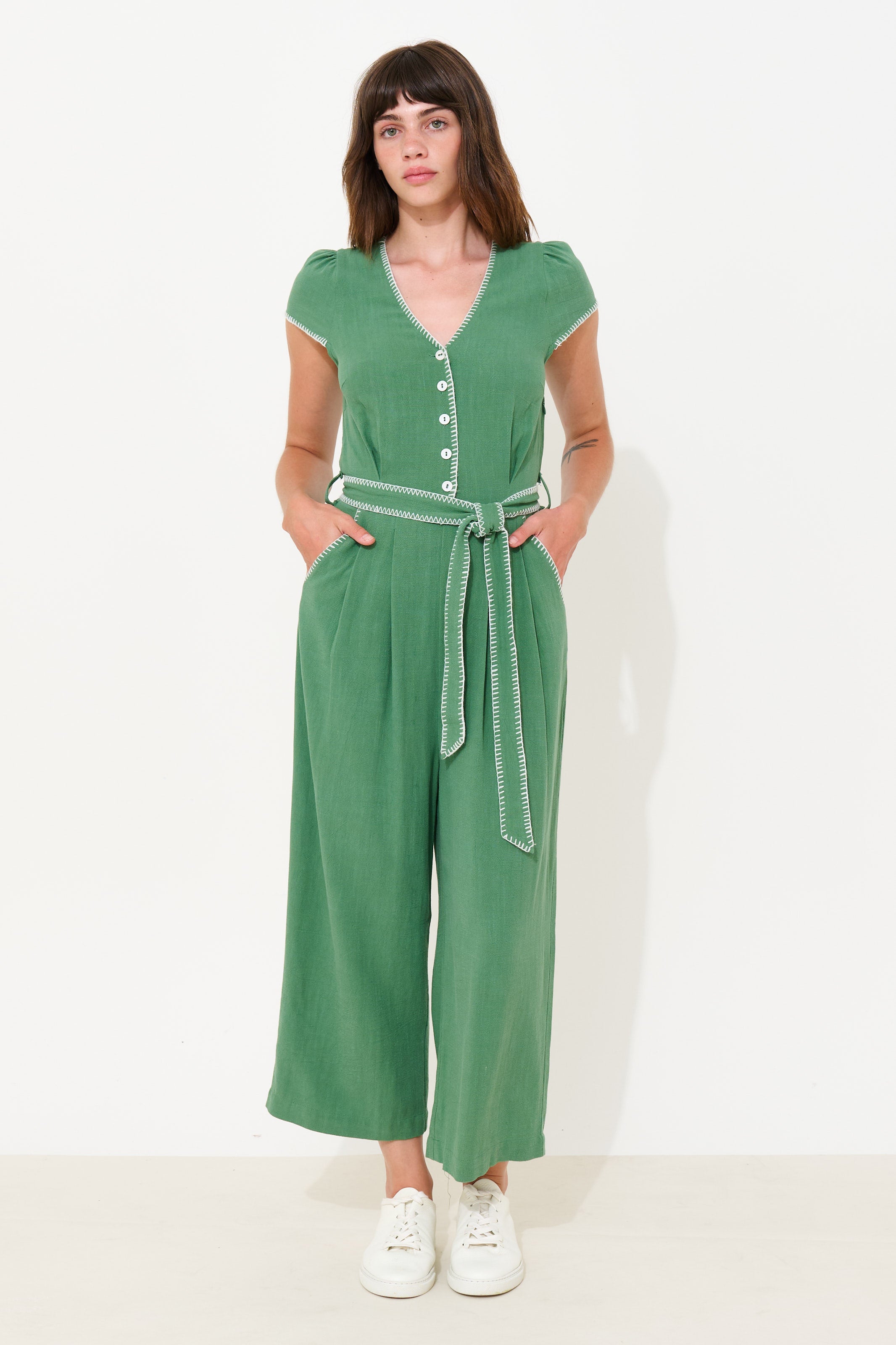A woman modeling a green short-sleeve jumpsuit with a buttoned V-neck, wide legs, and a matching tie belt, standing against a plain white studio background.