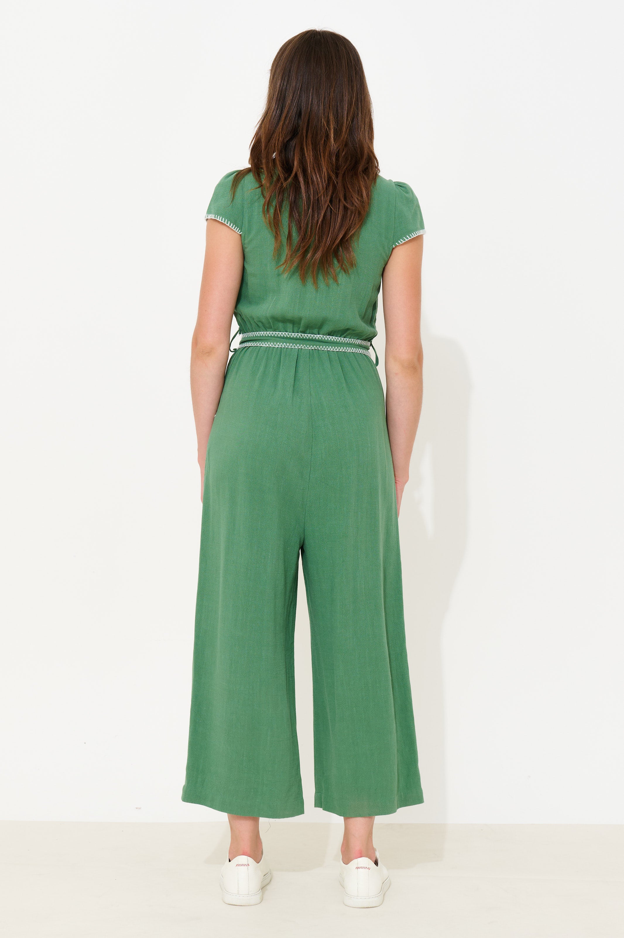 A woman viewed from the back wearing a green short-sleeve jumpsuit with a thin white waistband stripe, paired with white sneakers, against a plain white studio background.