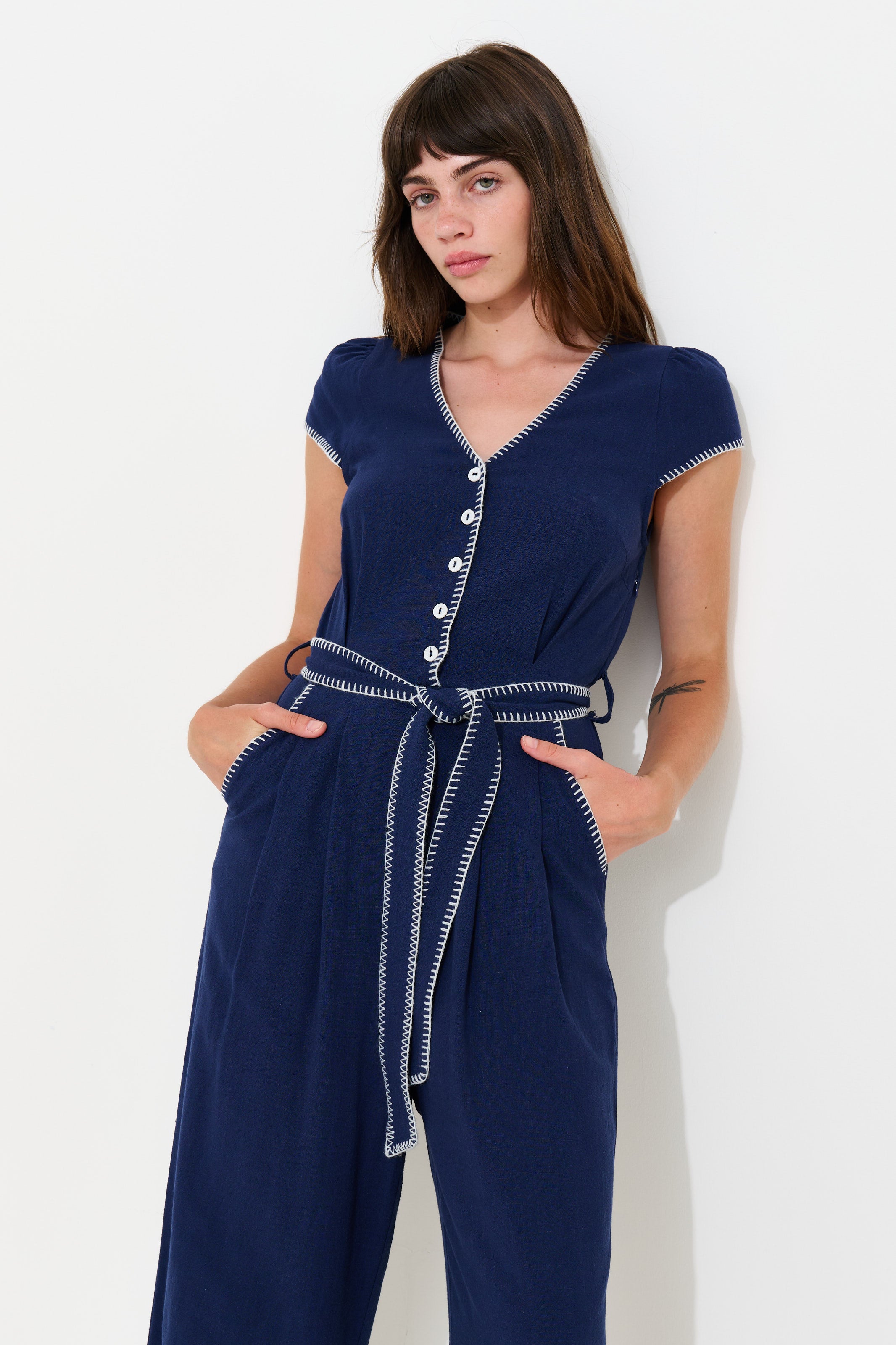 A woman modeling a navy blue jumpsuit with white contrast stitching, a buttoned V-neckline, and a matching tie belt, set against a plain white background.