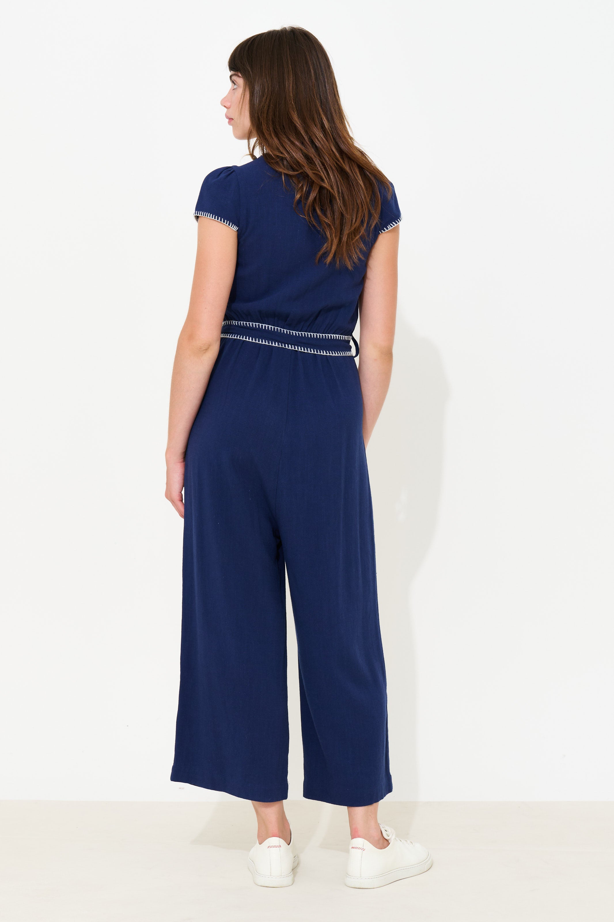 A woman wearing a navy blue jumpsuit with a thin silver chain detail at the waist, shown from the back against a plain white studio background.