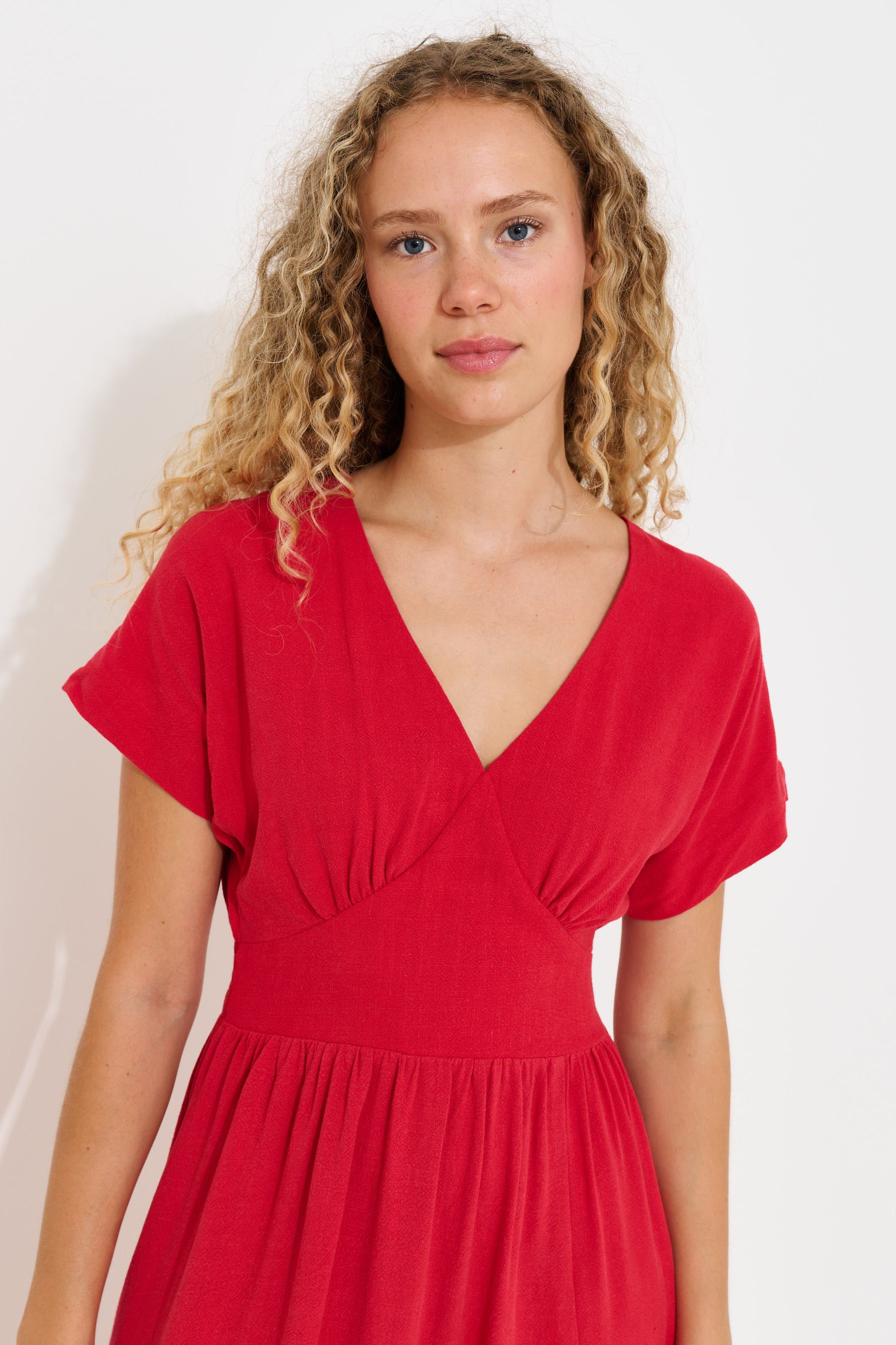 A young woman with curly blonde hair wearing a bright red short-sleeve dress with a V-neckline and gathered waist, photographed against a plain light background.