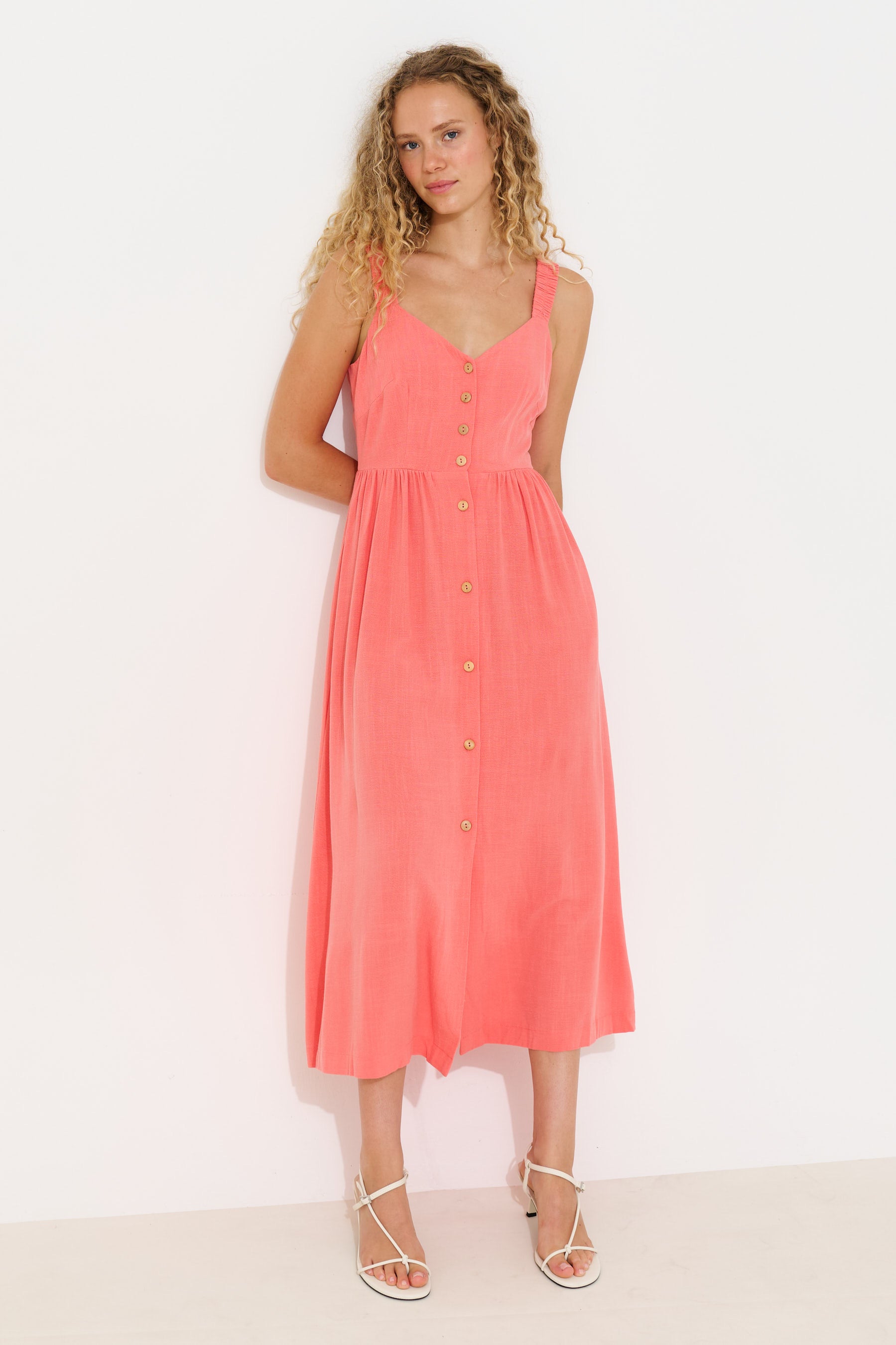 A woman modeling a coral pink sleeveless midi dress with a button-down front, photographed against a plain white studio background.