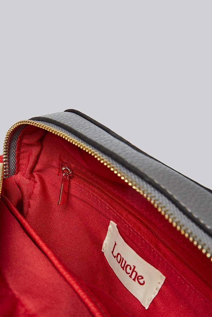 Louche Daisy camera bag detail — close-up of red leather tassel pull and gold-plated hardware against contrast panels.