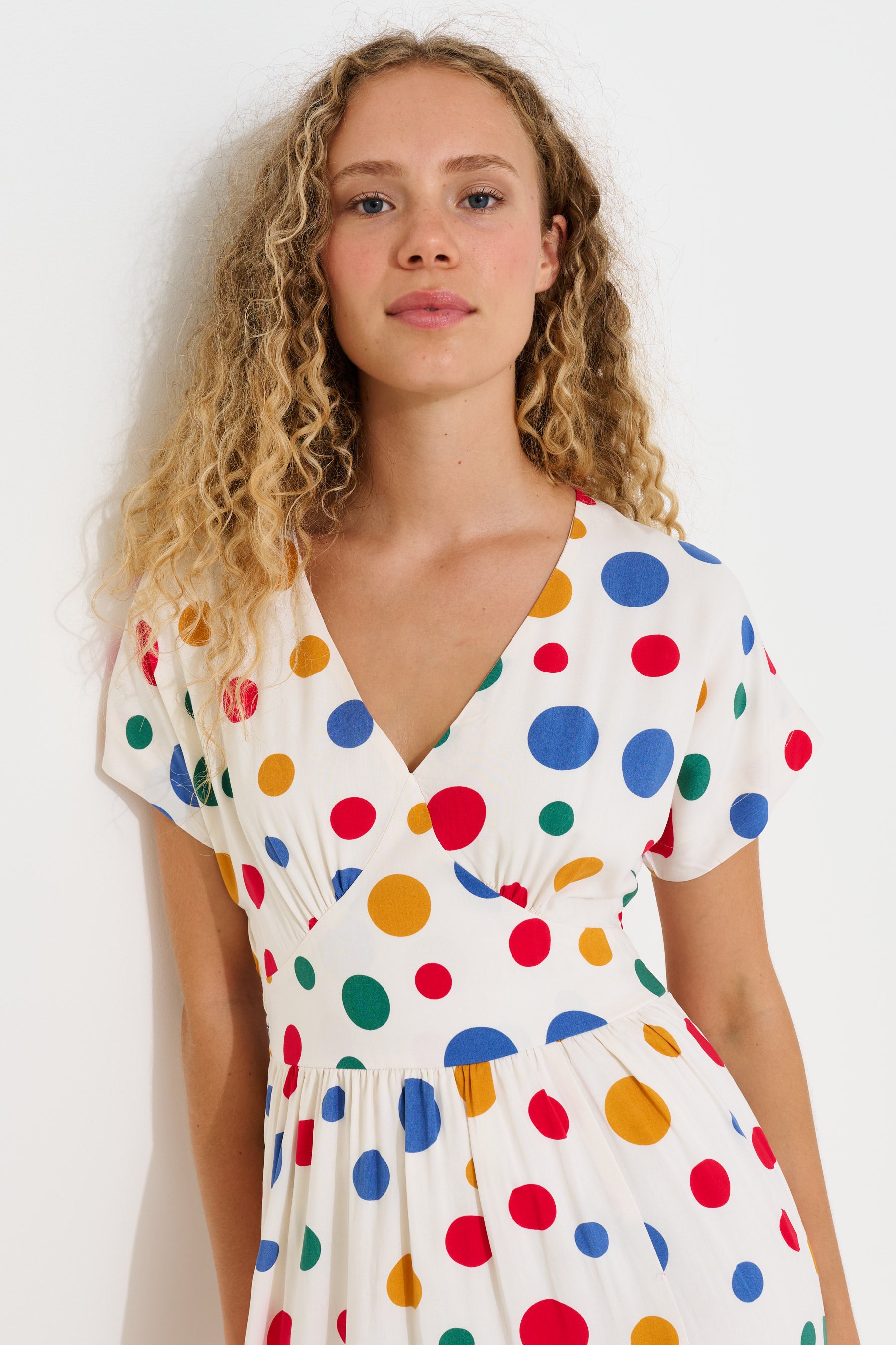 A woman with curly blonde hair wearing a white V-neck dress with a colorful polka dot pattern, photographed against a plain white background.
