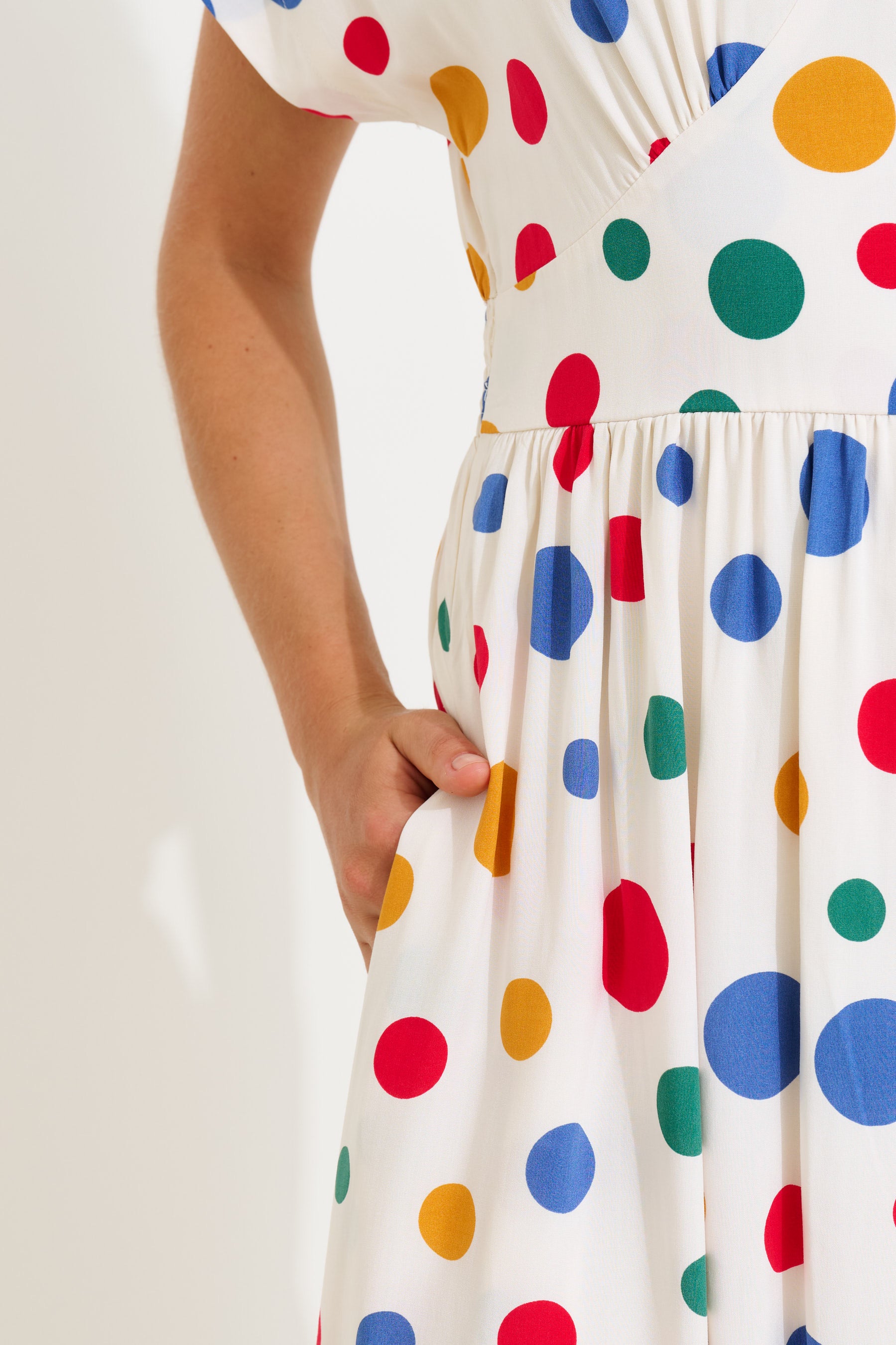Close-up of a person wearing a white dress with large colorful polka dots in red, blue, green, and yellow, with one hand tucked into a side pocket.